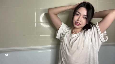 Video of a young Asian woman with straight, dark hair, wearing a white t-shirt, standing against a beige-tiled wall with a white lower section, arms raised, smiling.
