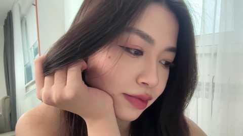 Video of an Asian woman with long black hair, light skin, and a slender physique, holding her hair, in a softly lit room with white curtains and a window.