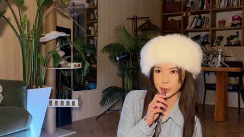 Video of a young Asian woman with long brown hair, wearing a fluffy white fur hat, licking an ice cream cone in a cozy, bookshelf-filled living room.