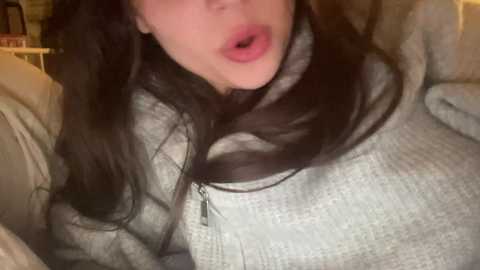 Media: A close-up video of a fair-skinned woman with long, dark hair, wearing a grey sweater, lips slightly parted, indoor setting, blurred background.