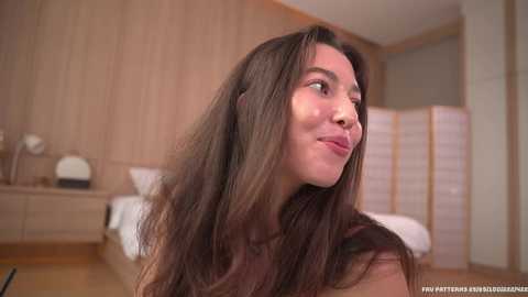 Media: Video of a young woman with long brown hair, fair skin, and a serene expression, standing in a modern bedroom with light wood walls, a bed, and a partition.