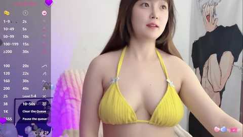 Video of a young East Asian woman with long brown hair, wearing a yellow bikini, standing in a room with a white wall and anime posters.