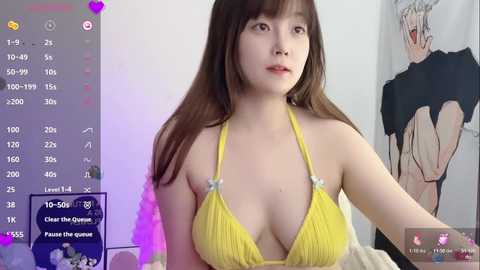 Video of an Asian woman with long brown hair, wearing a yellow halter bikini, sitting on a bed. Background features a wall with a poster of a cartoon character.