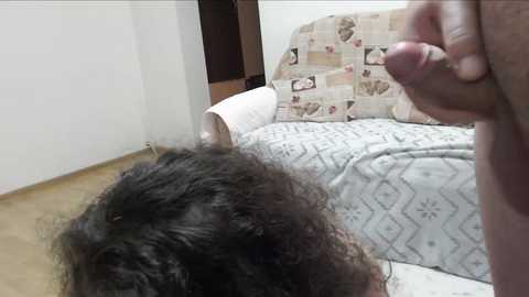 A video showing a naked, dark-skinned man with an erect penis, standing over a woman with dark curly hair, lying on a bed with a patterned quilt.