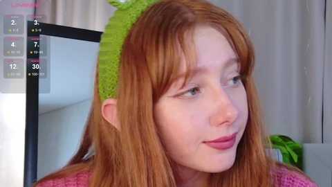 A video of a young Caucasian woman with long, straight, reddish-brown hair, wearing a green knitted headband with a frog design. She has fair skin, and is smiling slightly, with pink lipstick. The background is a blurred, indoor setting.