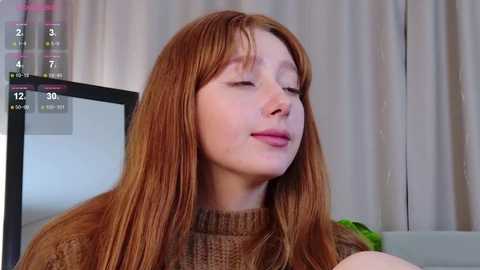 Video of a young Caucasian woman with long, straight, auburn hair, wearing a brown knitted sweater, eyes closed, smiling, in a room with beige curtains and a TV screen displaying a game.