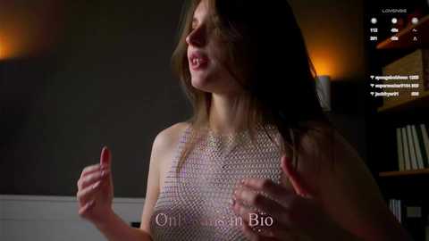 Video of a woman with long brown hair, wearing a sleeveless, patterned top, mid-conversation, indoors. Dark background with shelves and warm lighting.