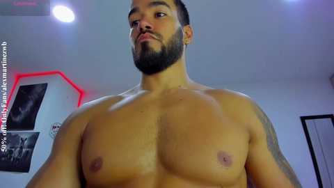 Video of a shirtless, muscular, light-skinned man with a trimmed beard and tattoos, standing indoors with a wall-mounted red light in the background.