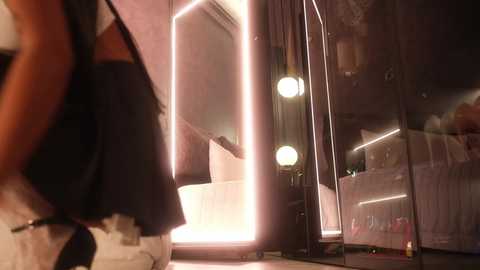Media: Video of a dimly lit bedroom with a large, glowing mirror reflecting a woman in a black dress. The room features a bed with white sheets and a stuffed toy, and soft lighting creates a cozy ambiance.