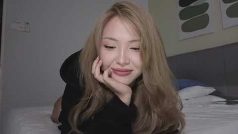 Video of a young Asian woman with long, wavy blonde hair, light skin, and pink lipstick, resting her chin on her hand, smiling softly. She wears a black top. Background includes a bed with white sheets, gray headboard, and modern art on the wall.