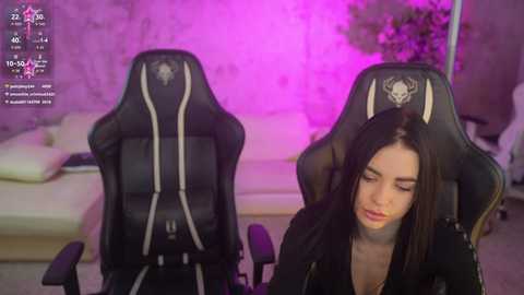 Video of a woman with long black hair wearing a black top, seated in a black gaming chair with skull design, in a modern room with purple lighting.