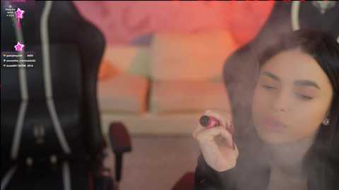 Video of a woman with long black hair, wearing a black leather jacket, applying makeup in a smoky room with a pink couch.