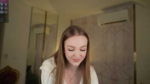 Video of a smiling young Caucasian woman with straight brown hair, wearing a white top, standing indoors in a modern room with a beige marble wall, air conditioner, and a TV.
