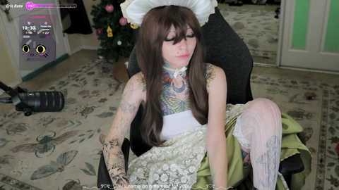 A video shows a fair-skinned woman with long brown hair in a maid costume, seated in a chair with a green dress, intricate floral tattoo sleeves, and a white lace headpiece.