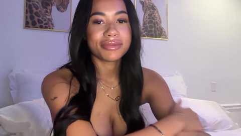Video of a smiling, light-skinned woman with long black hair, wearing a black bra, on a bed with white pillows and leopard-print wall art.