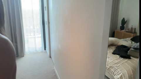 A video of a modern bedroom with a large mirror reflecting a cluttered bed and a potted plant. The room has light-colored walls, beige carpet, and sheer white curtains allowing soft natural light.