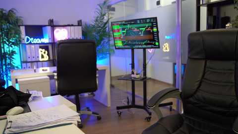 Video of a modern trading room with a black leather chair, computer screens displaying financial data, and a potted plant, illuminated by soft blue and purple lighting.