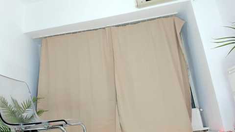 A video of a beige curtain partially drawn over a window, with a metal-framed chair and a fern plant visible in the foreground. The room is bright and minimalistic, with white walls.