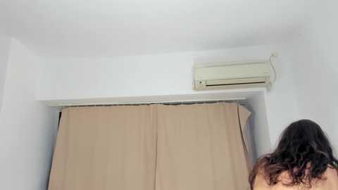 Video of a woman with long, wavy brown hair, viewed from behind, standing in a room with a white wall, beige curtains, and an air conditioning unit mounted on the wall.