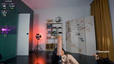 Video of a modern, dimly lit room with a large, erect penis on a table, a man with a tattooed arm, and a white door in the background.