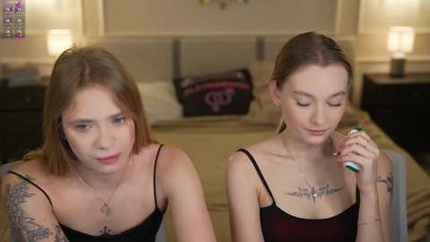 Video of two young women with light skin, straight brown hair, and black tank tops in a dimly-lit bedroom. One has a tattoo on her left arm, while the other holds a toothbrush, smiling.