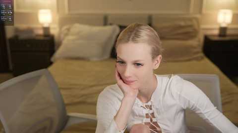 Media: Video of a blonde woman with fair skin, wearing a white top, resting her chin on her hand, seated at a table in a modern bedroom with beige bedding and lamps on nightstands.