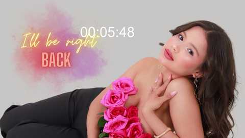 Video of an attractive Asian woman with long brown hair, wearing a black strapless dress adorned with a large pink rose, posing against a soft gradient background. Text reads, \"00:05:48 It'll be right back BACK.\