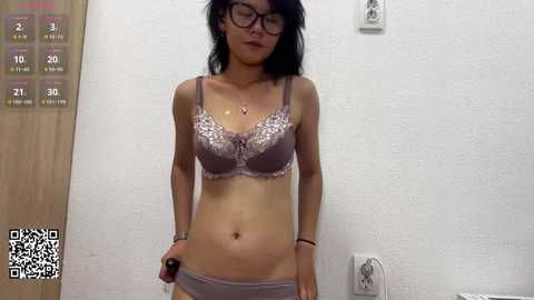 A video of a slender Asian woman with long black hair, wearing glasses, and lavender lingerie, standing in a white room with a wooden door and electrical outlets.