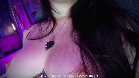 Video of a woman with long dark hair, fair skin, wearing a black lace bra, and a black pendant necklace. Background features purple and blue lighting, creating a sensual, intimate atmosphere.