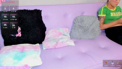 A video shows a woman with a tattooed arm, wearing a green t-shirt, sitting on a pastel purple couch. The background features colorful, tie-dye pillows and a black, fuzzy throw.