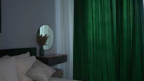 Video of a modern bedroom with a green curtain, white bedding, and a round mirror with a vase of dried flowers on a wooden nightstand.