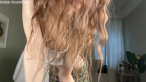 Video of a fair-skinned woman with long, wavy, light brown hair, seen from the side. She wears a leopard print top. Background features a window with white curtains, a plant, and a wooden table.