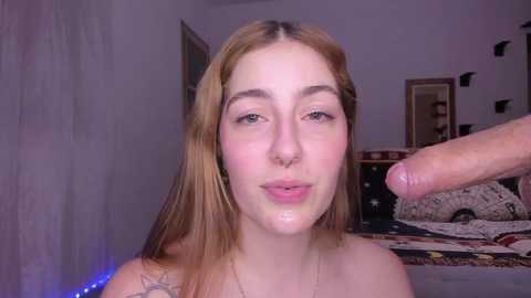 Media: Video of a young woman with light skin and straight, light brown hair, wearing a sleeveless top, kissing an erect penis in a dimly lit bedroom with a bed and white walls.