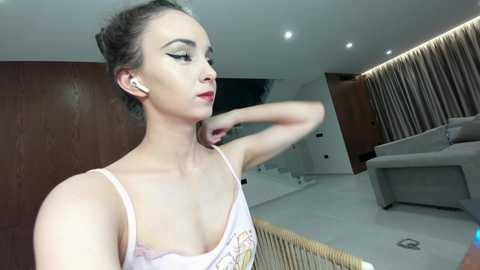 Video of a slender, light-skinned woman with dark hair in a bun, wearing a pastel-colored tank top, standing in a modern, minimalist living room with wooden paneling and white walls.