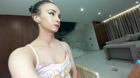 Video of a slender, light-skinned woman with dark hair styled in a bun, wearing a pink tank top with a cartoon graphic. She stands indoors, looking to the right, with a modern, minimalist living room in the background.