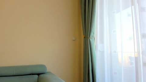 A video of a modern living room corner featuring a light blue couch against a pale beige wall. To the right, a large window with sheer white curtains and matching drapes is partially open, allowing soft natural light to filter in.