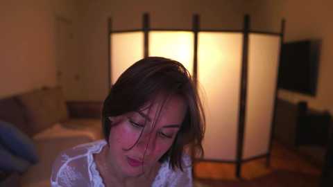 Media: Video of an Asian woman with straight, shoulder-length brown hair, wearing a white lace top, sitting in a dimly lit living room with a warm-toned lamp and a wooden floor.