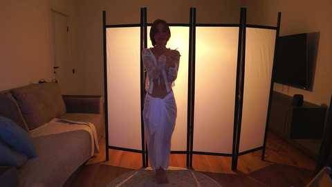 Video of a woman in a white dress, standing in a dimly-lit living room with beige furniture, a TV on the wall, and a partition screen.