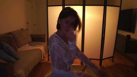 Media: Video of a woman with shoulder-length hair in a white lace dress, seated on a wooden floor in a dimly lit living room with a screen divider and beige couch.