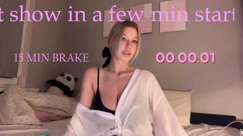 Video of a blonde woman with a medium build in a white robe, sitting on a bed in a cozy room with a panda toy and pink posters. Text overlays read \"15 MIN BRACE\" and \"0:00:01\".