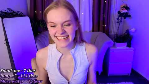 Video of a young, smiling woman with fair skin and shoulder-length blonde hair, wearing a white sleeveless top with a plunging neckline. She sits in a dimly lit room with purple lighting, surrounded by a computer monitor, a white dresser, and a decorative plant.