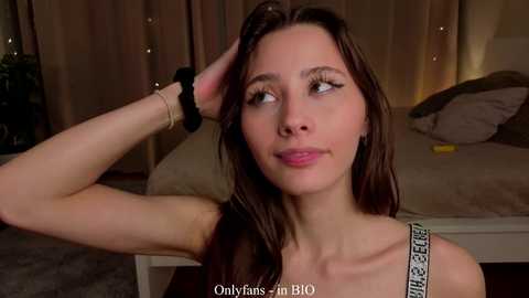 Video of a young woman with long brown hair, wearing a blue bra, standing in a dimly lit bedroom with beige curtains and a bed.