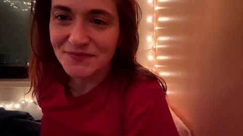 Video of a smiling woman with long, wavy, auburn hair, wearing a red shirt, standing in a warmly lit room with string lights and a window visible in the background.