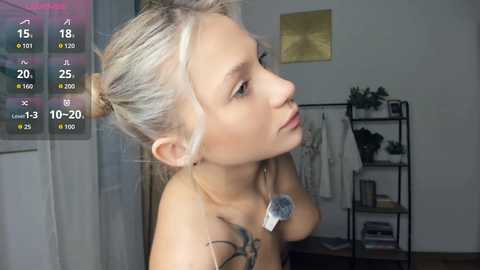Media: Video of a topless, pale-skinned woman with platinum blonde hair in a messy bun, wearing a dog tag necklace. She gazes pensively to the left, with a background featuring a modern, minimalist room with a black metal shelf, a potted plant, and a wall-mounted painting.