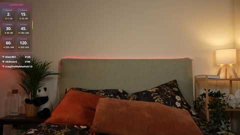 A video of a cozy bedroom with a green upholstered headboard, orange and floral-patterned pillows, a bedside table with a lamp, a potted plant, and a digital clock displaying 2:15.