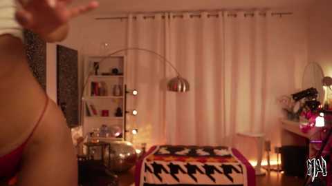 Video of a cozy, dimly-lit bedroom with a woman's bare shoulder visible on the left, a colorful blanket on a chair, and a desk with a mirror and candles in the background.