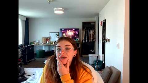 Video of a young woman with long brown hair and glasses, wearing a yellow sweater, sitting in a modern living room with a TV, cabinets, and a closet in the background.