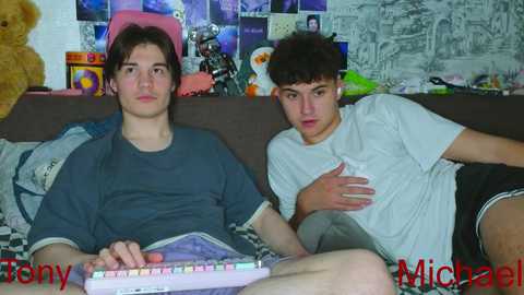 Video of two young men, one in a grey t-shirt and shorts, the other in a white t-shirt, sitting on a couch, playing a video game.
