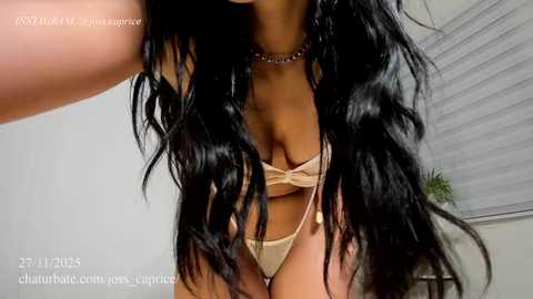 Video of a woman with long, wavy black hair, wearing a beige bra and matching panties, standing in a dimly lit room with blinds.