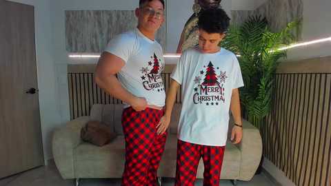 Video of two muscular men in white \"Merry Christmas\" T-shirts and red-and-black checkered pajama pants in a modern living room.